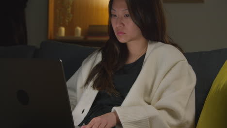 Woman-Spending-Evening-At-Home-Sitting-On-Sofa-With-Laptop-Computer-Looking-At-Social-Media-Streaming-Or-Scrolling-Online